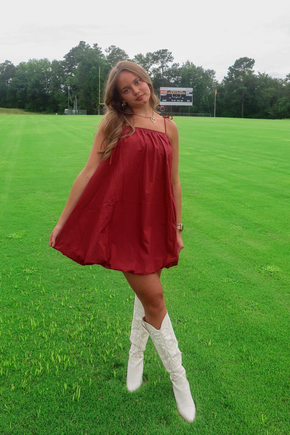 Woman in a red dress and white boots standing on a grassy field with trees in the background