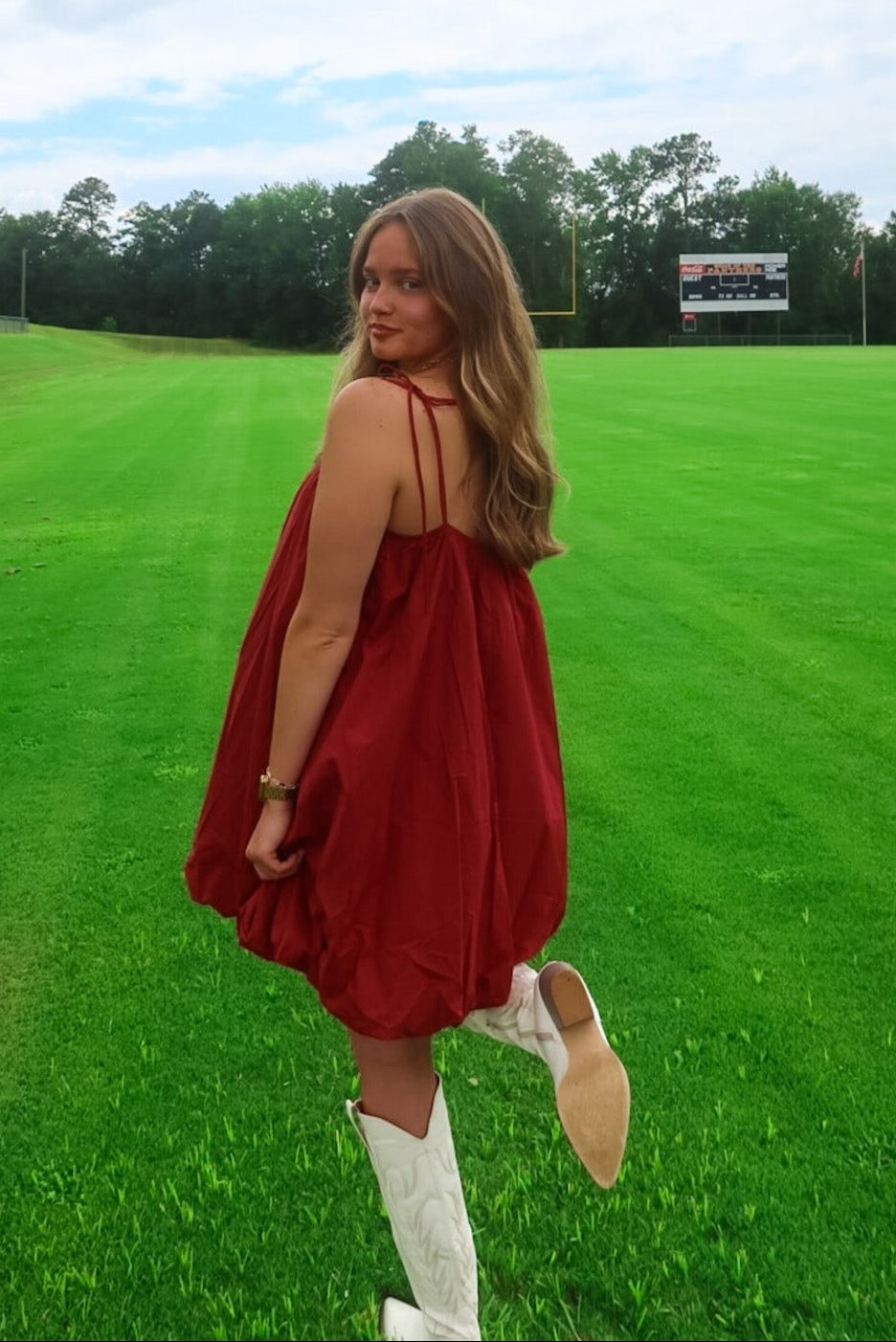 Woman in a red dress standing on a grassy field with trees and a building in the background.
