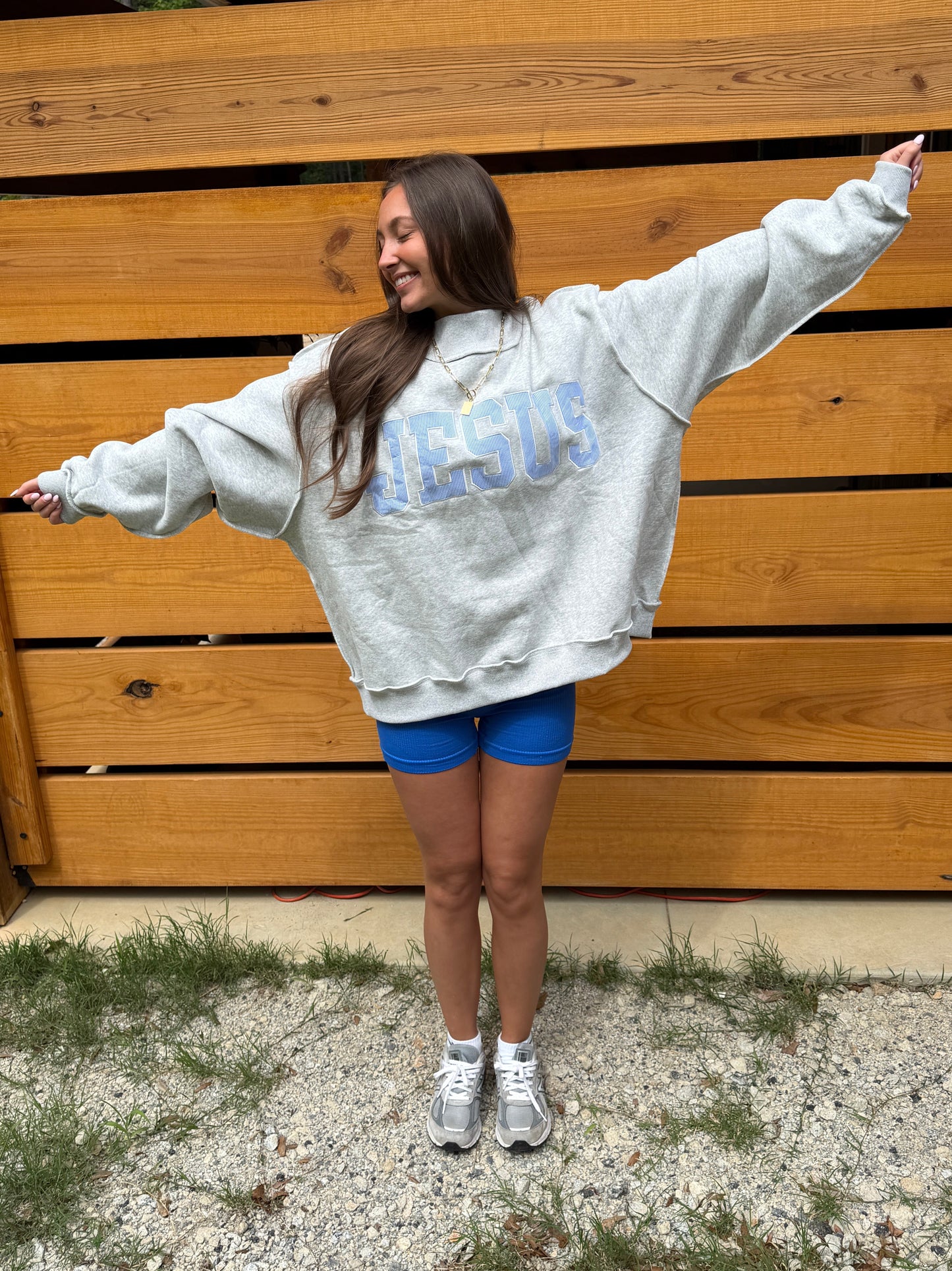 Person wearing a gray sweatshirt with 'JESUS' printed on it, standing in front of a wooden wall.