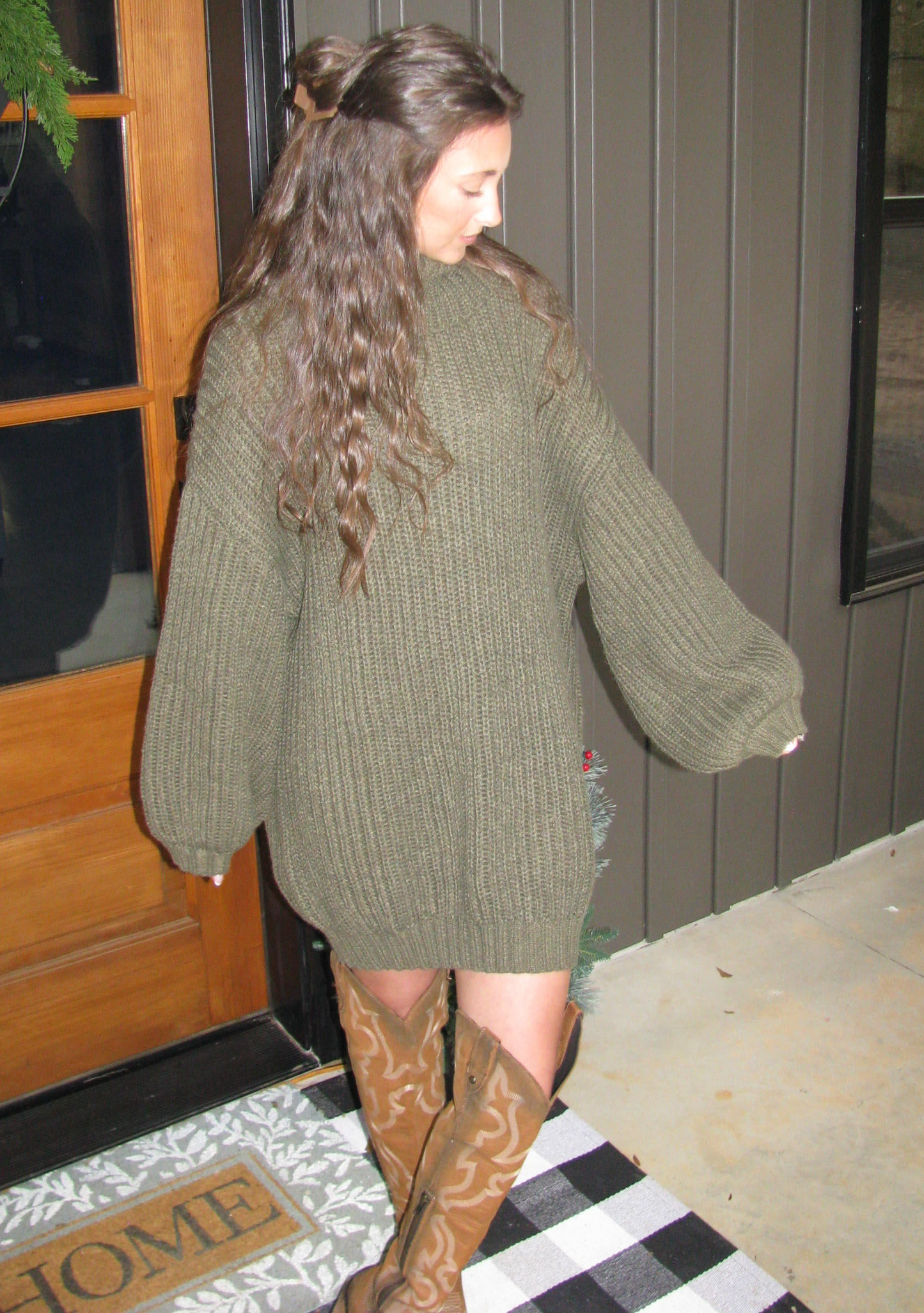 Person wearing a green sweater standing in front of a door with a plant and decorative item.