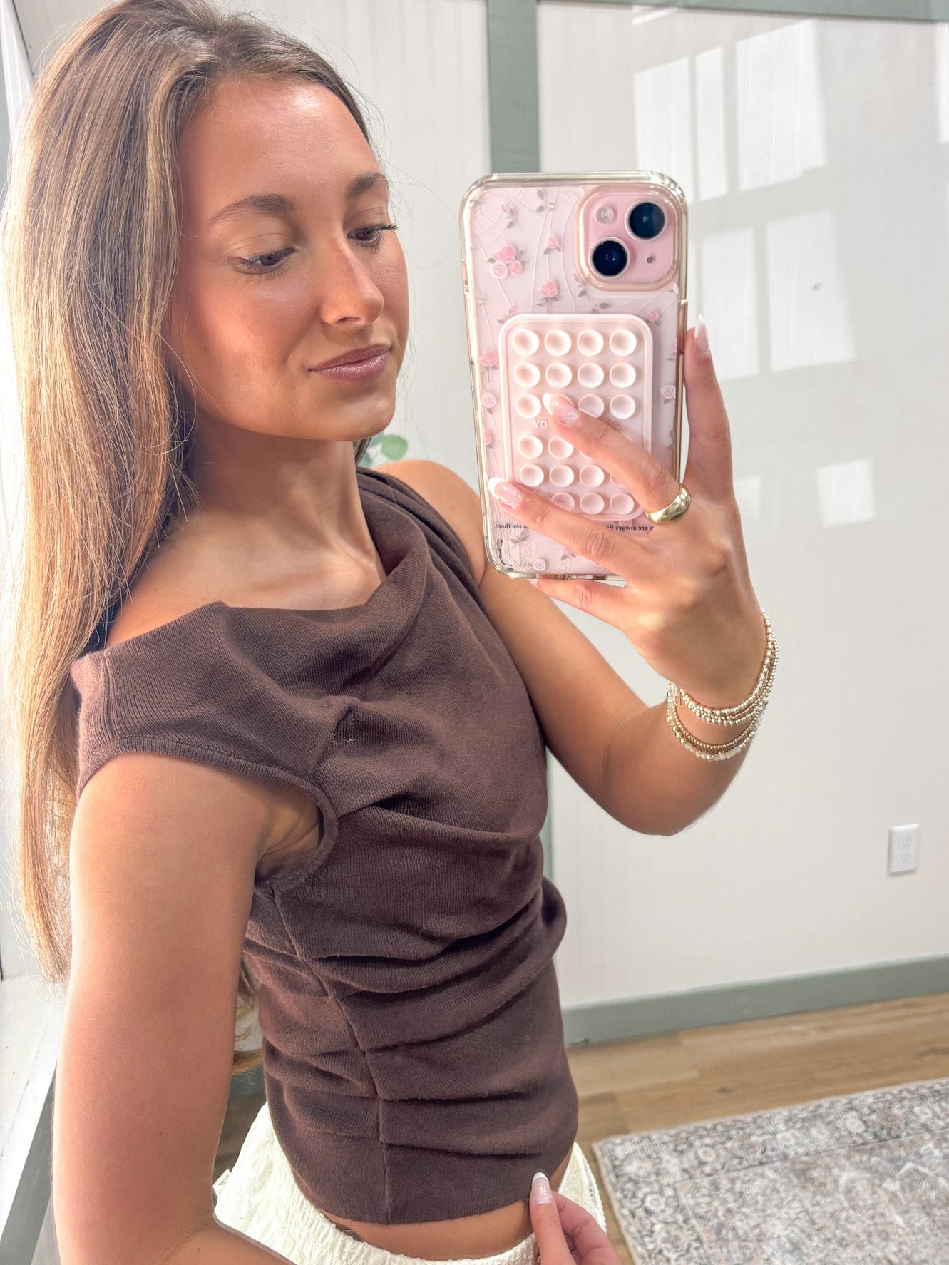 Woman taking a mirror selfie wearing a brown top in a room with a white wall and wooden floor.