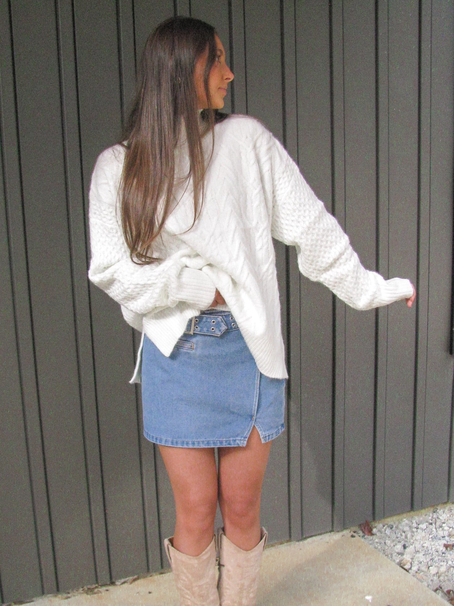 Woman wearing a white sweater, denim skirt, and beige boots standing against a gray wall.