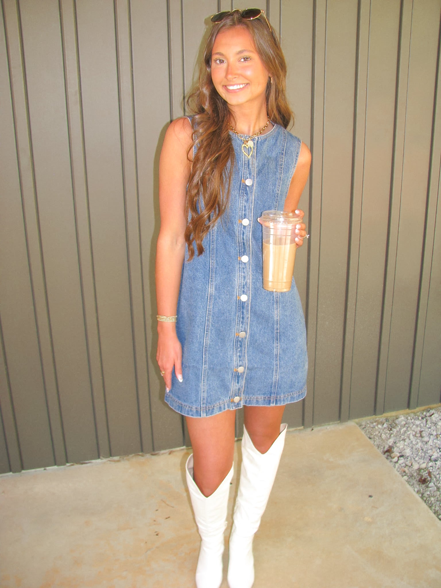 Woman in a denim dress and white boots holding a drink against a neutral background