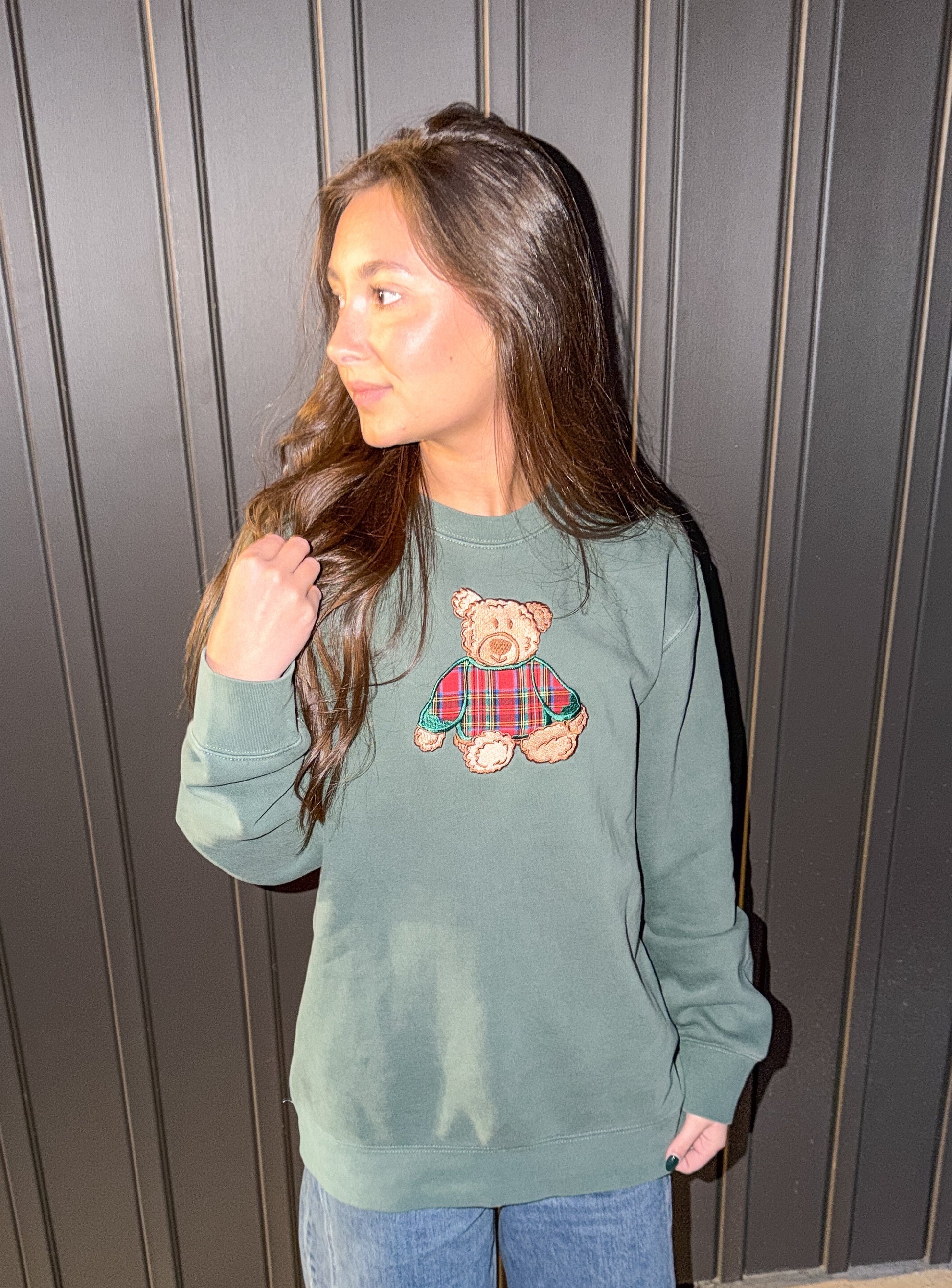 Woman wearing a green sweatshirt with a teddy bear design against a dark wooden panel background