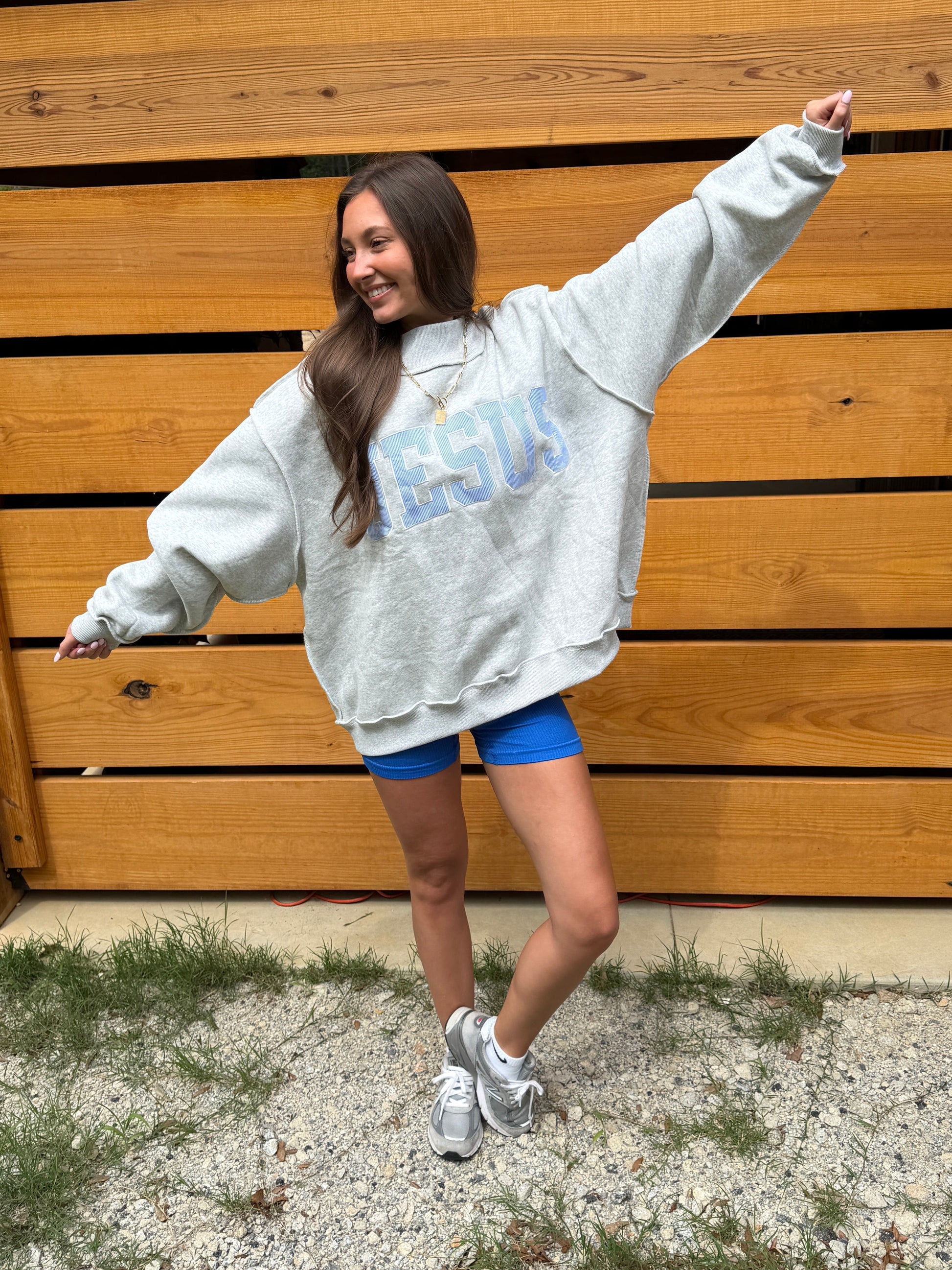 Person wearing a gray sweatshirt with blue text saying ‘JESUS’ blue shorts, and white sneakers, standing in front of a wooden fence.