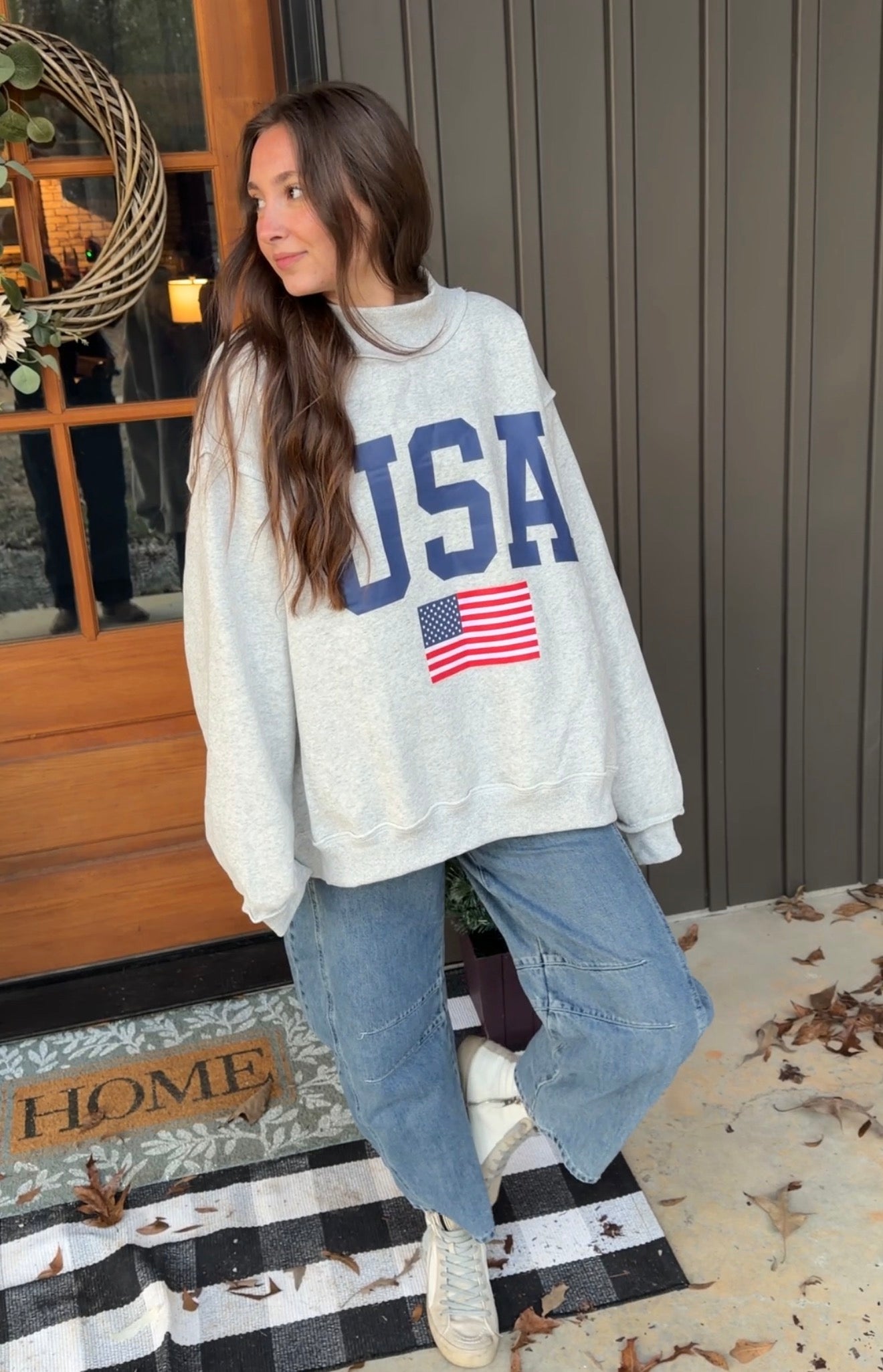 Woman wearing a white sweatshirt with 'USA' and an American flag design, standing on a porch.