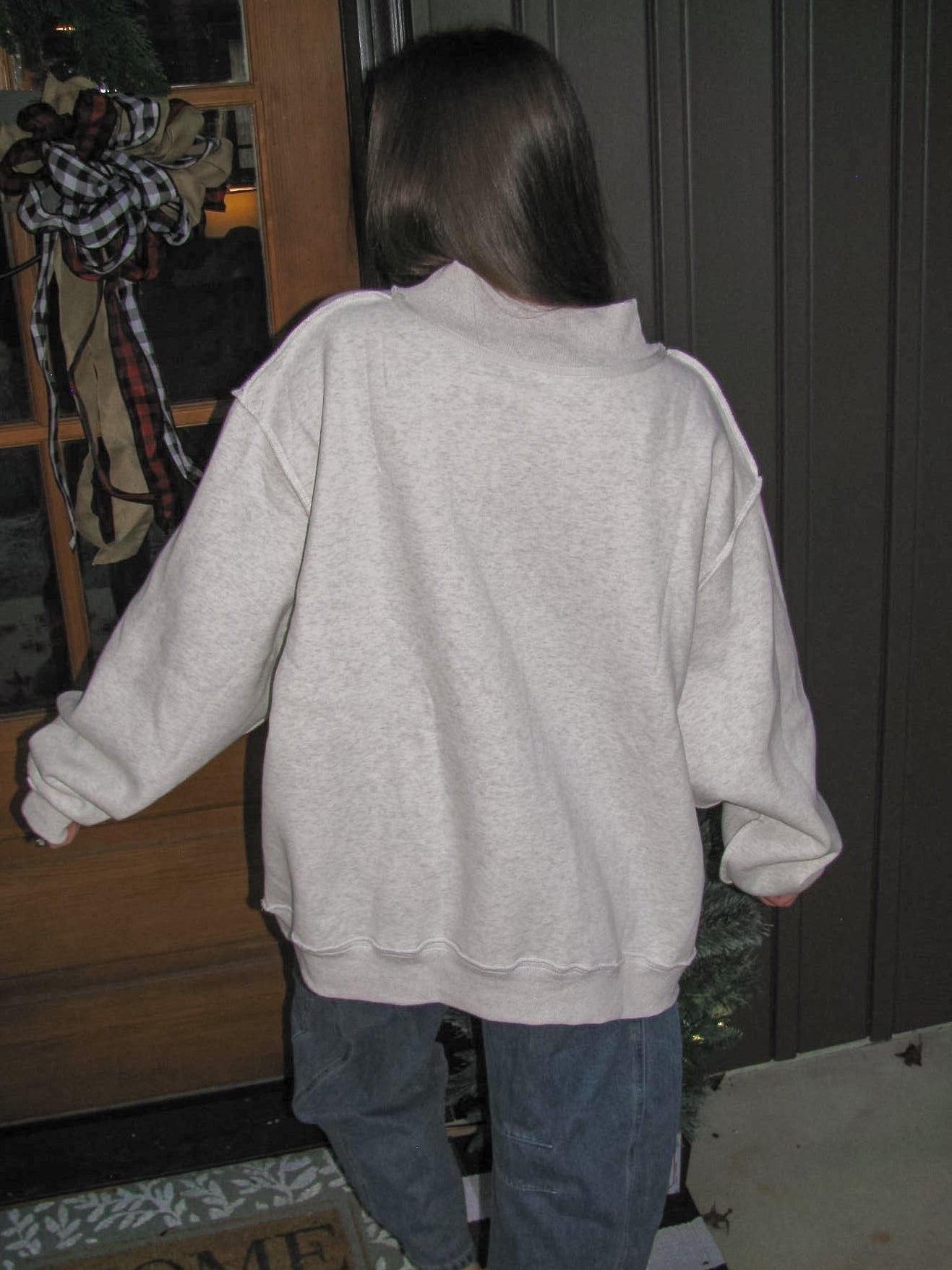 Person wearing a light gray sweatshirt and blue jeans standing on a checkered floor with a wooden door and coat rack in the background.
