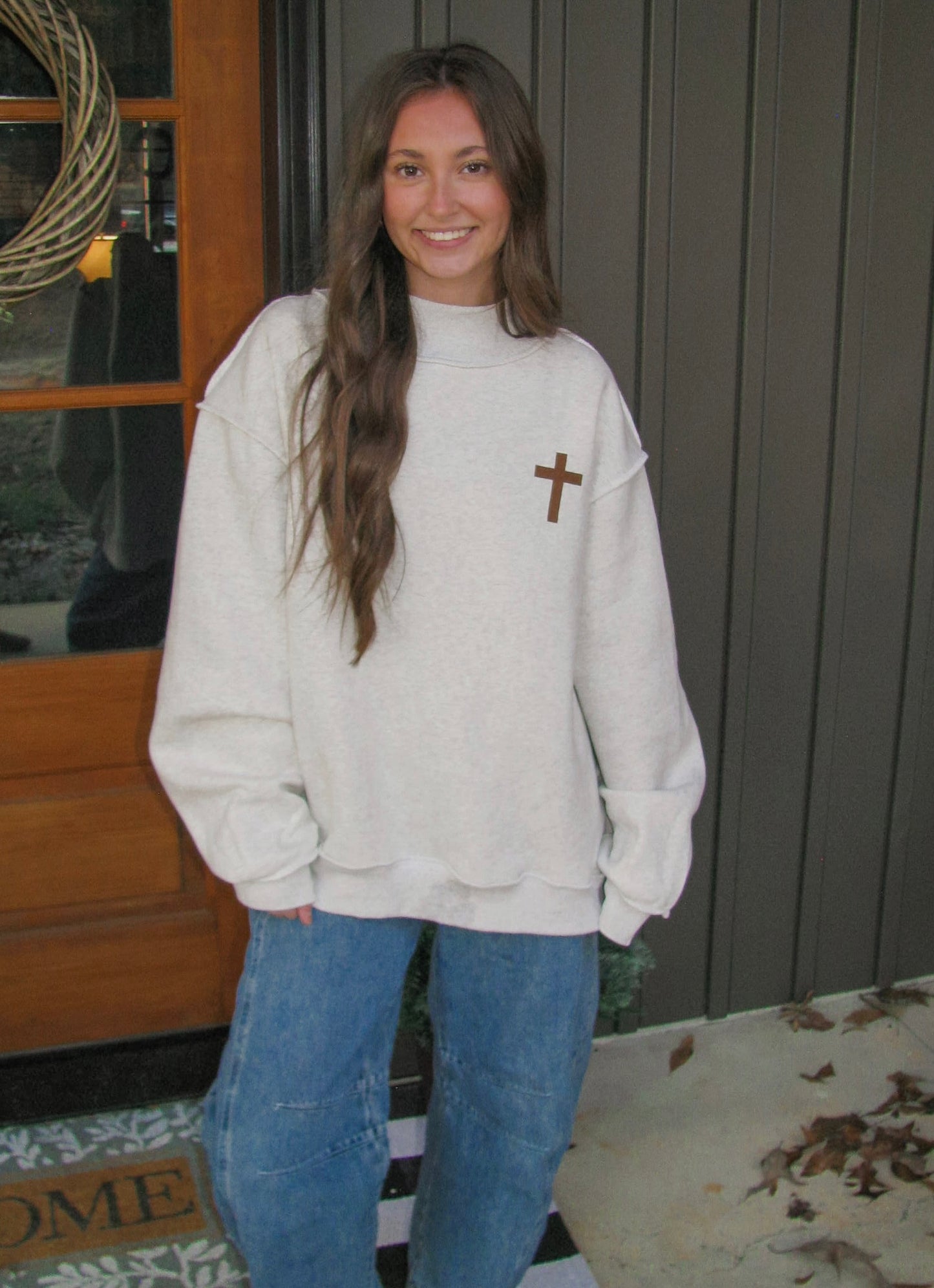 Person wearing a light-colored sweatshirt with a cross design and blue jeans, standing in front of a wooden door.
