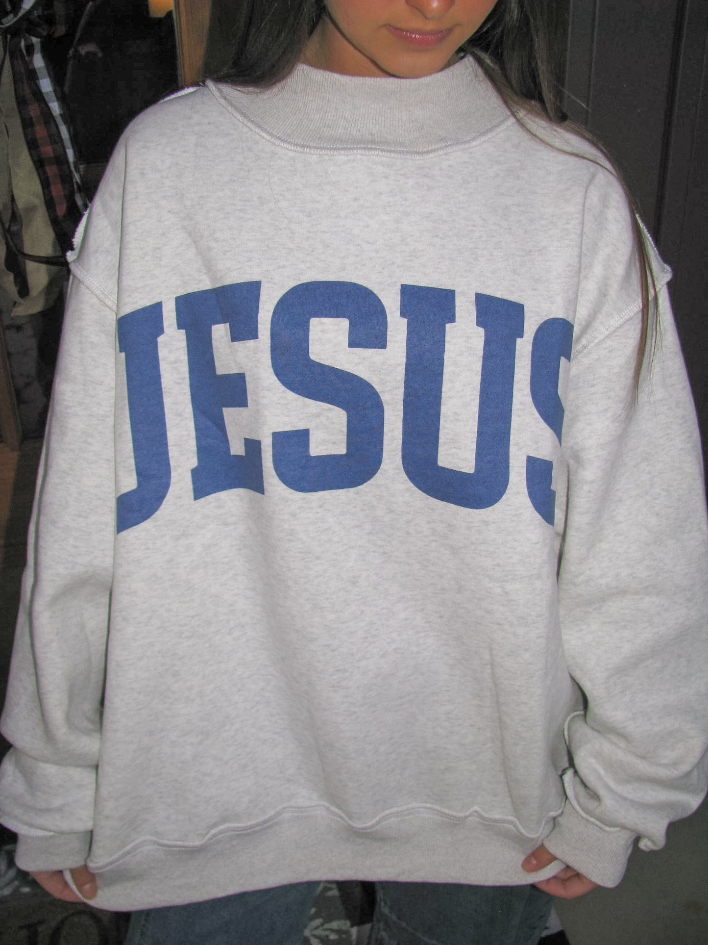 woman showing off gray sweatshirt with 'JESUS' printed on it.