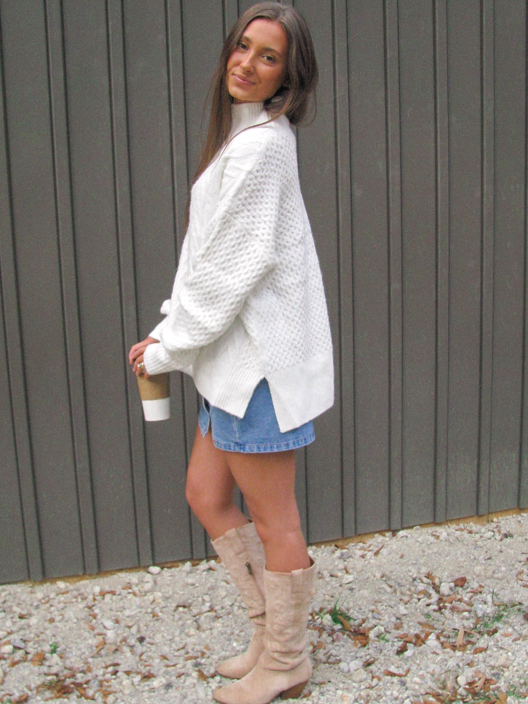 Woman wearing a white sweater, denim shorts, and beige boots against a gray wall.