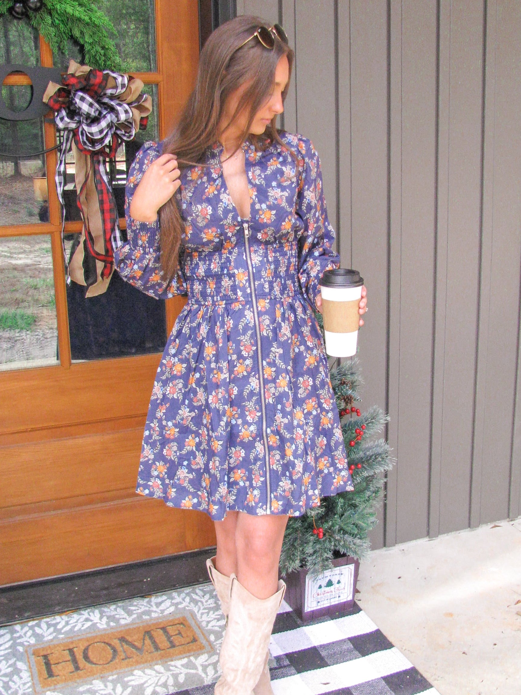 Woman in a blue floral dress holding a coffee cup on a porch with a Christmas tree.
