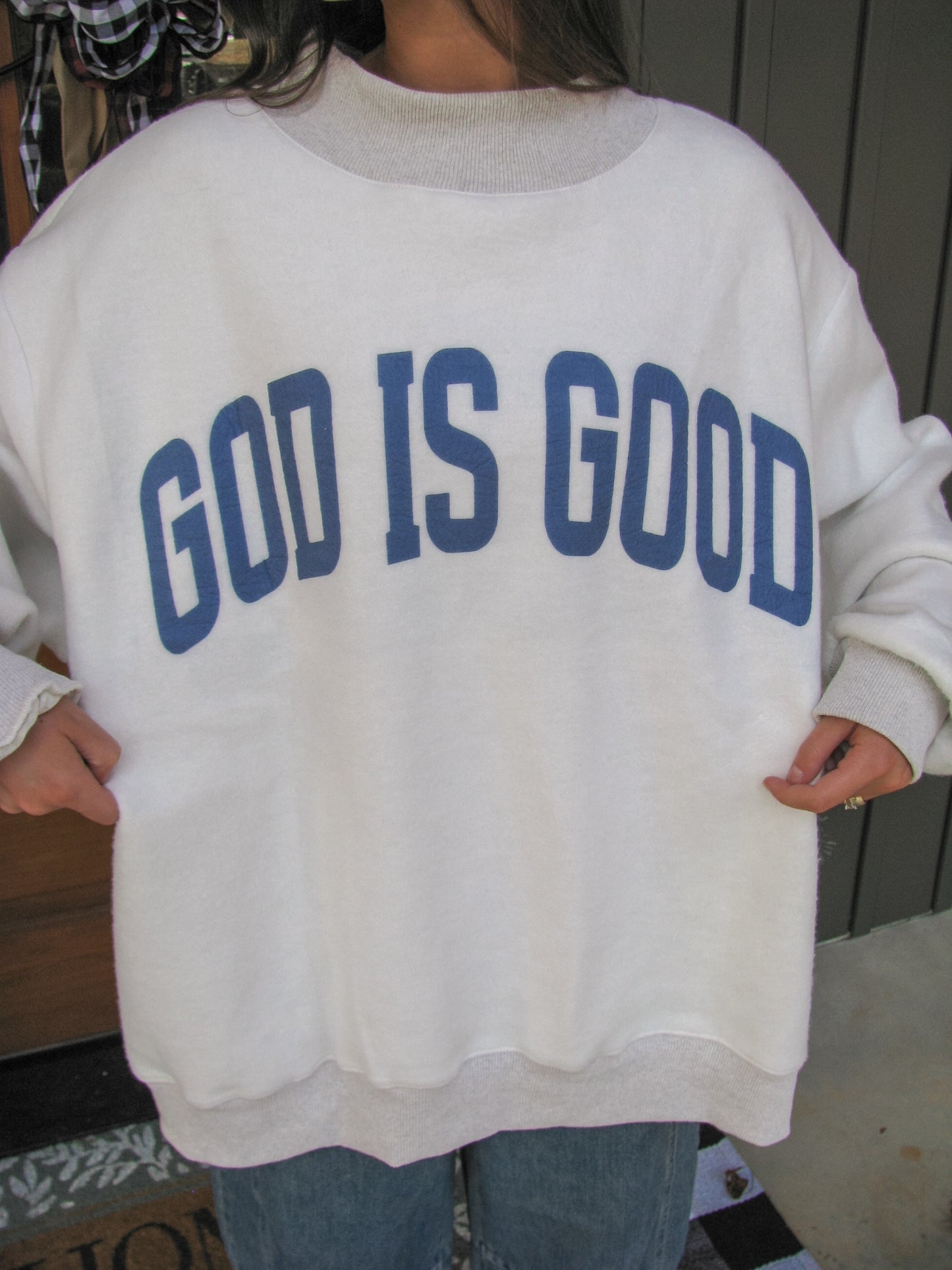 White sweatshirt with 'GOD IS GOOD' text in blue on a person indoors.