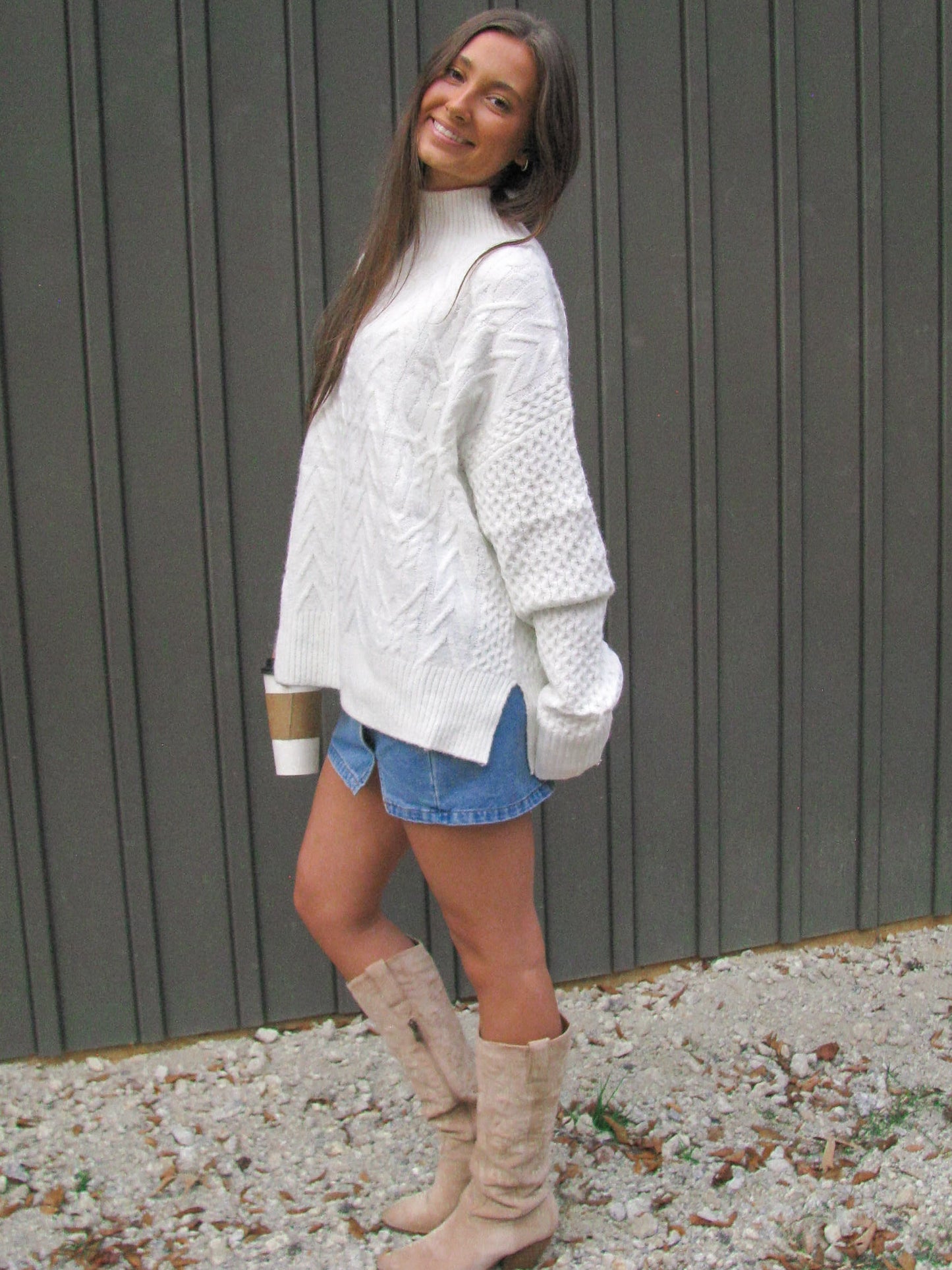 Woman wearing a white cable knit sweater, denim shorts, and beige knee-high boots standing against a gray wall.