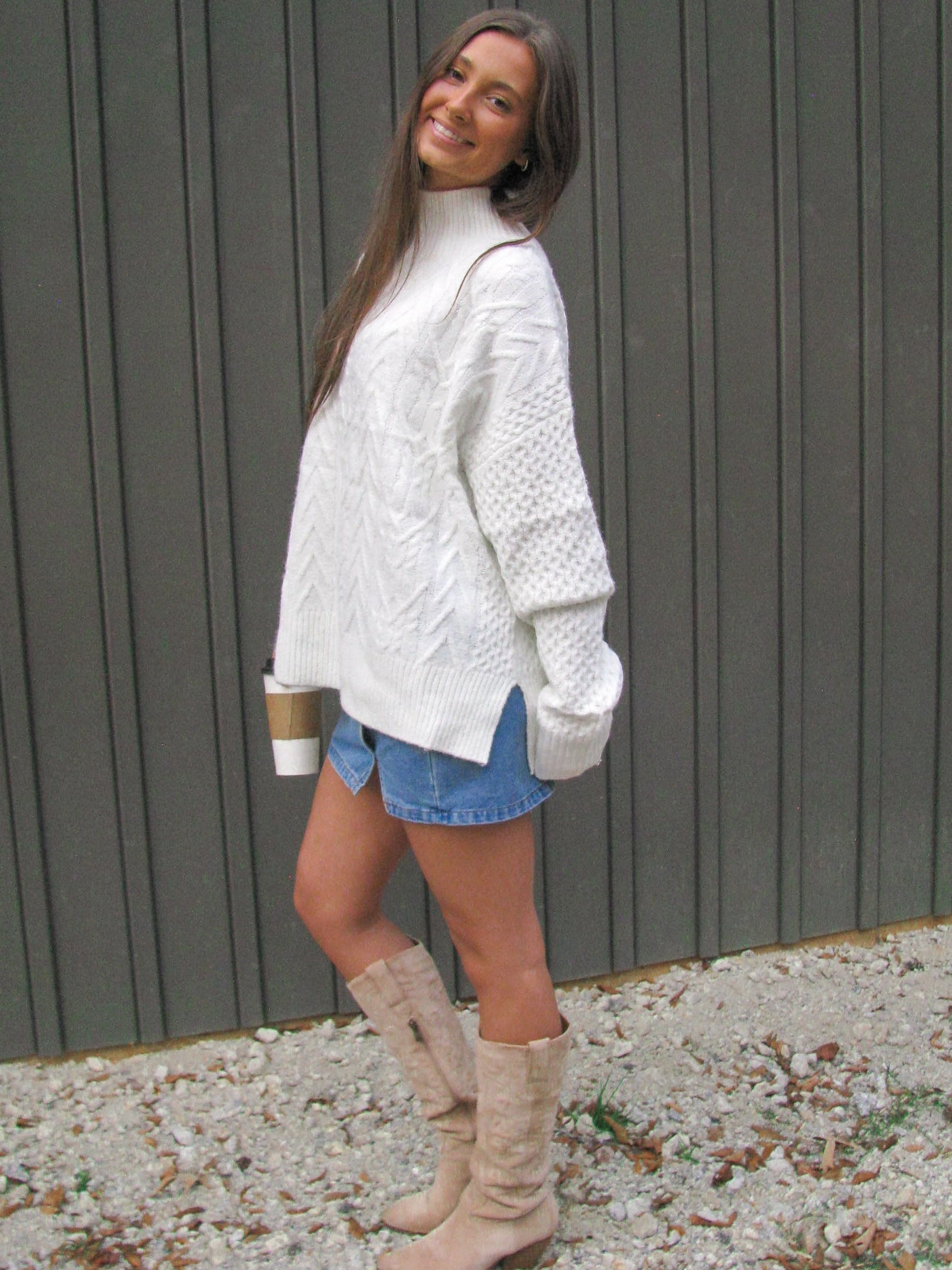 Woman wearing a white cable knit sweater, denim shorts, and beige knee-high boots standing against a gray wall.