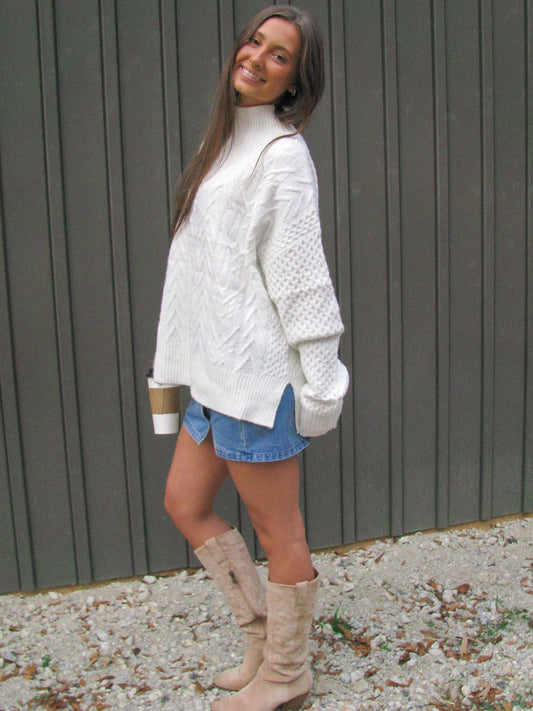 Woman wearing a white cable knit sweater, denim shorts, and beige knee-high boots standing against a gray wall.