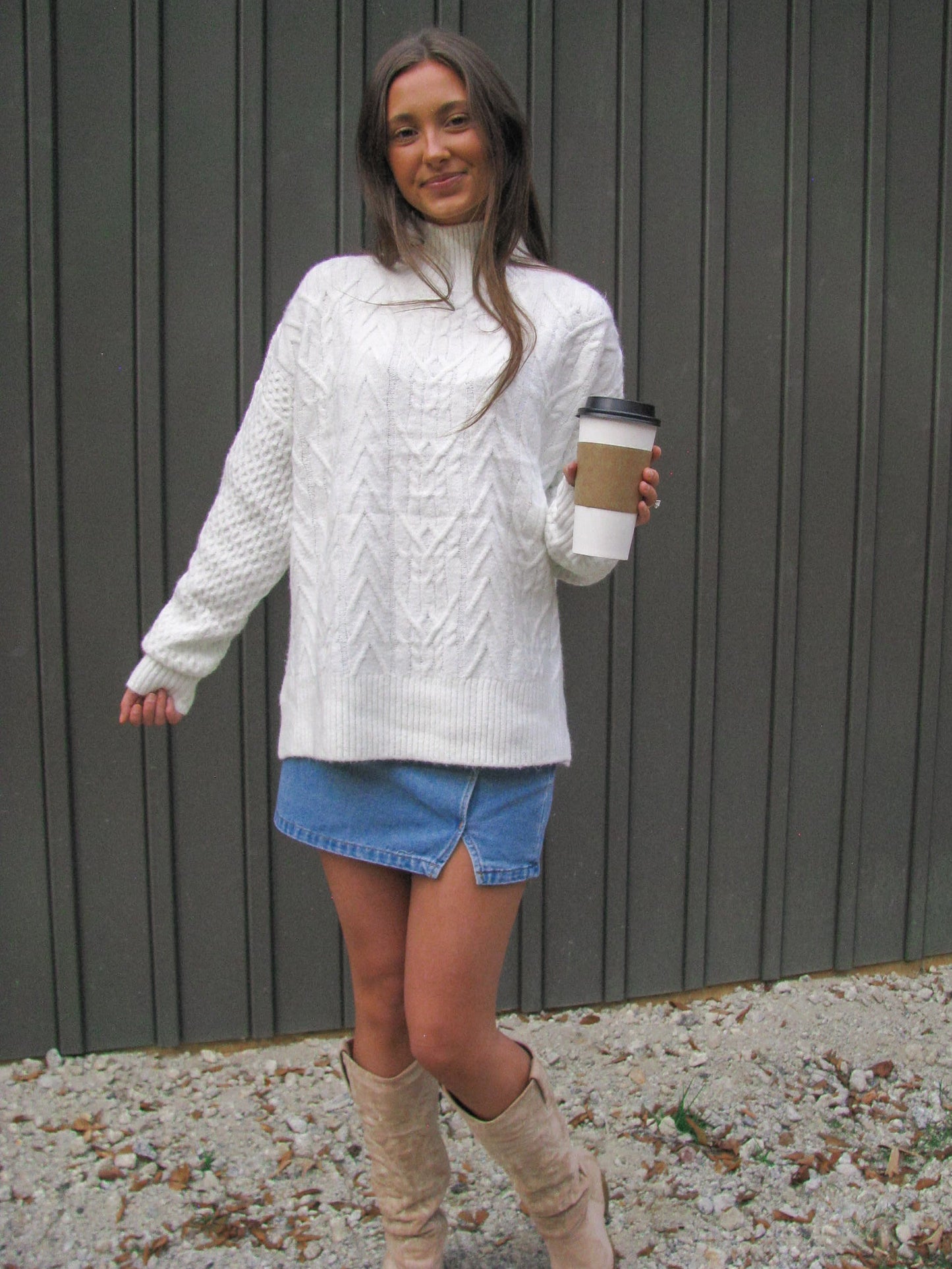 Woman wearing a white cable knit sweater, blue denim shorts, and beige knee-high boots, holding a coffee cup.